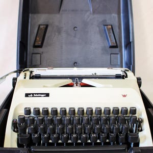 May include: Cream-colored Scheidegger typewriter with black keys and a black carrying case. The typewriter has a red ribbon and a small crown emblem. The case is open, revealing the typewriter inside.