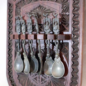 Puede incluir: Un juego de ocho cucharas de peltre con mangos ornamentados, exhibidas en un soporte de pared de madera con intrincados tallados. El soporte es de color marrón oscuro y tiene un borde festoneado.