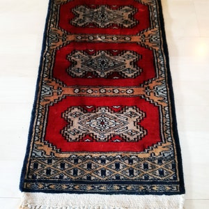 Peut inclure: Un tapis rectangulaire tissé à la main avec un champ rouge vif et des motifs géométriques complexes en beige, noir et or. Le tapis présente une bordure bleu foncé et des franges crème. Le tapis mesure environ 91 cm de long.