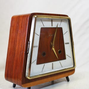 May include: A vintage wooden clock with a rectangular face and a gold-tone frame. The clock face is white with black markers and gold hands. The clock has a dark brown wooden body and sits on four short black legs.