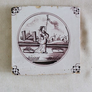 Può includere: Una piastrella di ceramica marrone e bianca con un design circolare. Il design presenta una persona con una lunga veste che tiene un bastone, in piedi in un paesaggio con una recinzione e alberi. La piastrella ha un bordo decorativo.