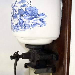 以下が含まれることがあります： 壁掛け式のアンティークコーヒーグラインダー。白いセラミック容器には、青と白のオランダの風車のシーンが描かれています。グラインダーは、黒い金属製のメカニズム、木製のハンドル、透明なガラス製の収集ジャーを備えています。濃い色の木材の裏板に取り付けられています。