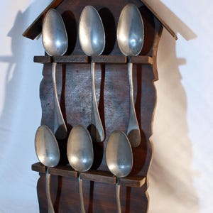 Puede incluir: Un soporte de cucharas de madera con forma de casa con techo a dos aguas, que contiene seis cucharas de plata. El soporte es de color marrón oscuro y está montado en una pared blanca.