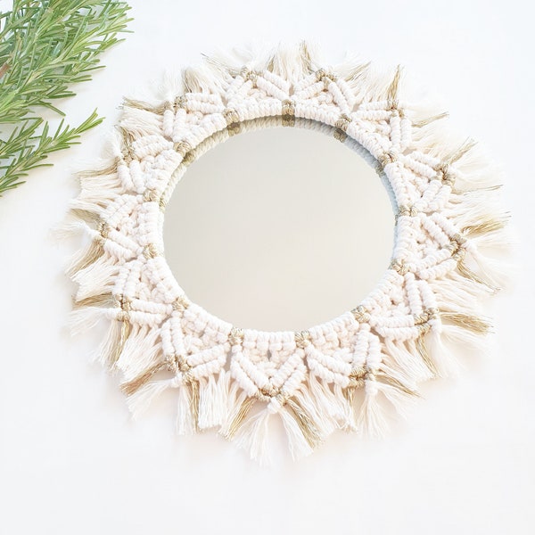 Può includere: Uno specchio in macramé bianco e oro con uno specchio rotondo e un bordo a frangia.