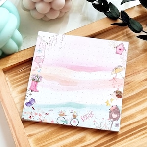 Könnte beinhalten: Ein quadratischer Notizblock mit Frühlingsmotiv. Der Notizblock zeigt eine Aquarellillustration mit rosa, blauen und weißen Farbtönen. Das Design umfasst ein Fahrrad mit Blumen, einen rosa Stiefel mit Blumen und das Wort "Spring".