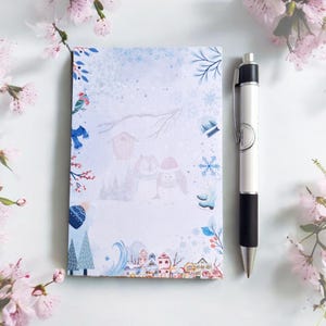Könnte beinhalten: Ein Notizblock zum Thema Winter mit einem Stift. Der Notizblock zeigt Illustrationen von Eulen, Häusern und Schneeflocken in Blau-, Weiß- und Rottönen. Ein weißer Stift mit schwarzem Griff liegt neben dem Notizblock.