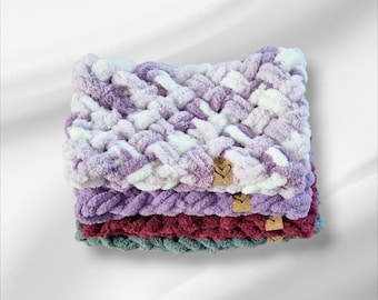 May include: A stack of four chunky knit scarves in shades of purple, white, burgundy, and teal. Each scarf has a woven texture and a small, heart-shaped tag. The scarves are stacked on top of each other.