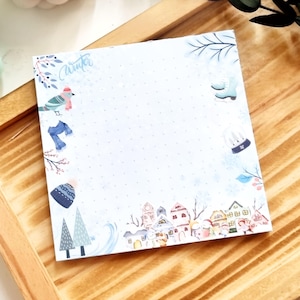 Könnte beinhalten: Quadratischer Notizblock mit Winterthema. Der Notizblock hat einen hellblauen Hintergrund mit einem gepunkteten Muster. In der oberen linken Ecke steht das Wort "Winter" in Schreibschrift. Er ist mit Winterillustrationen wie Vogel, Schal, Mütze und Häusern verziert.
