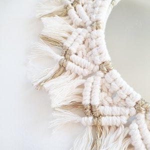 May include: A white macrame wall hanging with gold accents. The macrame is in the shape of a circle and has a fringe border.