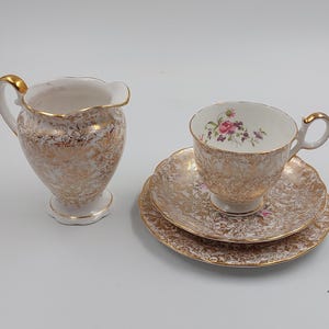 May include: A vintage teacup and saucer set with a matching creamer. The set features a white background with gold filigree and floral accents. The teacup has a gold handle and rim, and the saucer has a scalloped edge.