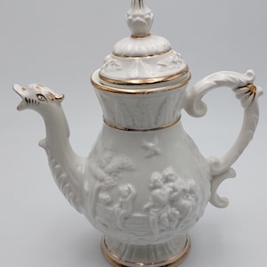 Puede incluir: Tetera de porcelana blanca con ribete dorado y un pico en forma de dragón. La tetera presenta un diseño en relieve de personas y árboles.