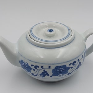 May include: White ceramic teapot with a blue floral pattern and matching lid. The teapot has a curved spout and handle. The lid features a small blue flower design in the centre and a blue ring around the edge.
