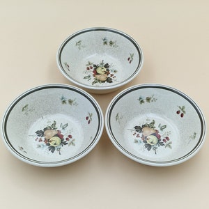 May include: Three white ceramic bowls with a green rim and a floral design featuring fruit and berries. The bowls are on a light beige background.