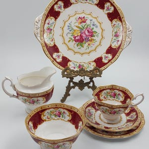 May include: A vintage tea set with a burgundy and gold floral design. The set includes a serving plate, creamer, bowl, teacup, and saucer. The plate is supported by a decorative metal stand. The teacup and saucer are stacked.
