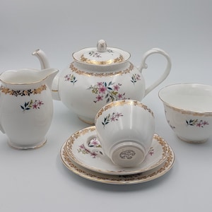 Könnte beinhalten: Ein weißes Teeservice mit goldenen Verzierungen und Blumendesigns. Das Set beinhaltet eine Teekanne, eine Milchkännchen, eine Zuckerdose, eine Tasse und eine Untertasse.
