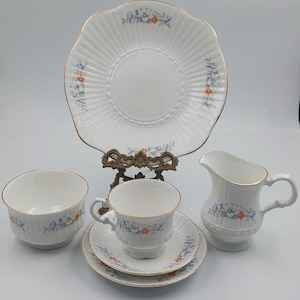 May include: A white porcelain tea set with a floral design. The set includes a large plate, a creamer, a teacup with saucer, and a small bowl. The edges are trimmed with gold. The floral pattern includes blue and orange flowers.