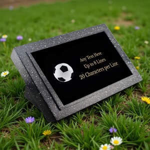 May include: A black and white football graphic is featured on a black plaque. The plaque is set in a speckled grey frame, with text that reads "Any Text Here, Up to 8 Lines, 20 Characters per Line." The plaque is displayed on a bed of green grass.