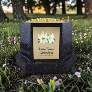 Puede incluir: Una urna conmemorativa negra y dorada con un diseño floral y el texto "In Loving Memory of Grandpa". La urna está situada en una zona de césped con flores silvestres y árboles al fondo.