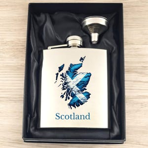 Könnte beinhalten: Eine Edelstahl-Taschenflasche mit silberner Oberfläche und einem blau-weißen schottischen Flaggen-Design. Die Flasche befindet sich in einer schwarzen Samtbox mit einem silbernen Trichter. Die Flasche trägt den Schriftzug "Scotland" auf der Vorderseite.