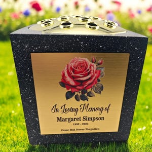 Puede incluir: Jarrón conmemorativo de granito negro con una placa dorada que presenta una rosa roja y la inscripción "En memoria cariñosa de Margaret Simpson 1952 - 2024 Se fue pero nunca será olvidada".