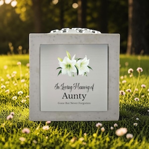 May include: A square, light gray memorial urn with a silver plaque featuring white lilies and the text "In Loving Memory of Aunty, Gone But Never Forgotten." The urn is set on a bed of green grass.