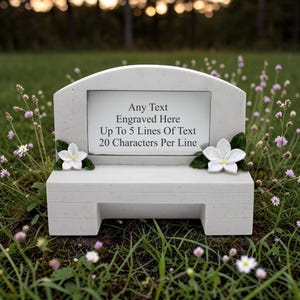 May include: A light grey memorial bench with space for engraved text. The bench features two white flower accents and is set in grass with wildflowers. Text on the bench reads: "Any Text Engraved Here Up To 5 Lines Of Text 20 Characters Per Line."