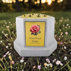 May include: A memorial vase with a gold plaque featuring a red rose and the inscription "In Loving Memory of Gran." The vase is light gray with a yellow top and sits in a grassy area with small white flowers.