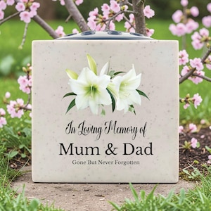 Op de afbeelding: Een vierkante, crèmekleurige herdenkingsvaas met een bloemenontwerp en de tekst "In Loving Memory of Mum & Dad, Gone But Never Forgotten." De vaas heeft een donkere bovenkant met gaten voor bloemstelen. Witte lelies en groene bladeren zijn gedrukt.