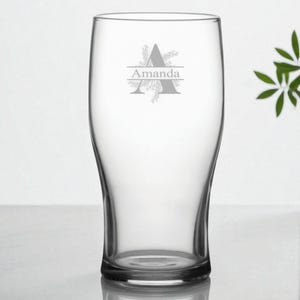 Könnte beinhalten: Ein klares, personalisiertes Bierglas mit dem Namen "Amanda" und einem dekorativen Anfangsbuchstaben "A" auf der Vorderseite. Das Glas hat eine klassische Form und eignet sich für Bier oder andere Getränke. Eine kleine Topfpflanze ist im Hintergrund.