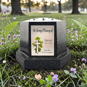 Könnte beinhalten: Schwarz-graue Gedenkurne mit dekorativer Oberseite, platziert in einer grasbewachsenen Fläche mit Blumen. Die Urne hat eine Metallplakette mit der Aufschrift "In Loving Memory of David 1945-2024 Gone But Never Forgotten" und ein Kreuzdesign.