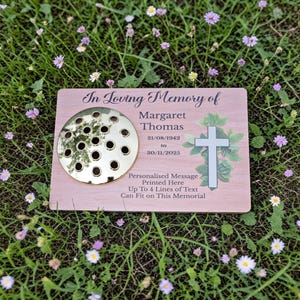 May include: A wooden memorial plaque with the text "In Loving Memory of Margaret Thomas" and dates. It features a gold-colored floral frog and a cross with foliage. The plaque is surrounded by green grass and small flowers.