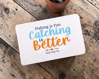 May include: A rectangular white metal tin featuring the text Fishing is Fun Catching is Better in blue and orange, with three small fish illustrations and the text Dad's Bait Tin printed in black at the bottom of the lid.