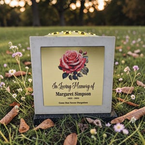 May include: A memorial urn with a gold plaque featuring a red rose illustration. The plaque reads "In Loving Memory of Margaret Simpson 1952-2024 Gone But Never Forgotten." The urn is white with a decorative top and sits on a black base.