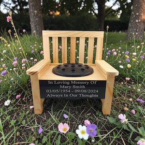 May include: A miniature wooden chair with a black floral foam insert. The chair features a black plaque inscribed with "In Loving Memory Of Mary Smith 03/05/1954 - 09/08/2024 Always In Our Thoughts". Set outdoors, surrounded by grass and flowers.