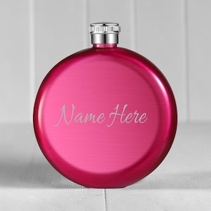 May include: A round, hot pink metal flask with a brushed finish and a silver-coloured screw-top cap. The front of the flask features the engraved text "Name Here" in a cursive script font, centred on the circular body of the container.