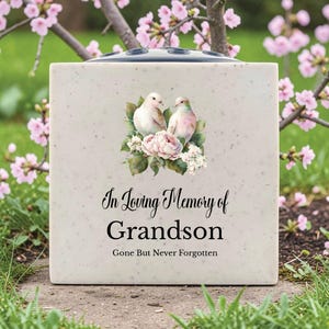 Op de afbeelding: Een vierkante, gespikkelde, gebroken witte herdenkingsvaas met een bloemen- en vogelontwerp. De vaas heeft de tekst "In Loving Memory of Grandson, Gone But Never Forgotten". De bovenkant heeft verschillende gaten voor bloemstelen.