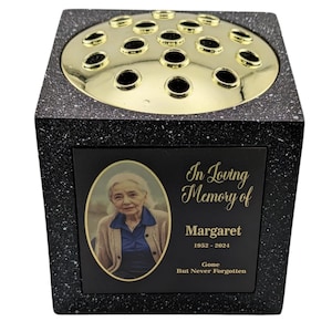 May include: A black and speckled memorial vase with a gold-colored floral grid. The vase features a photo of a woman and the inscription "In Loving Memory of Margaret 1952-2024 Gone But Never Forgotten."