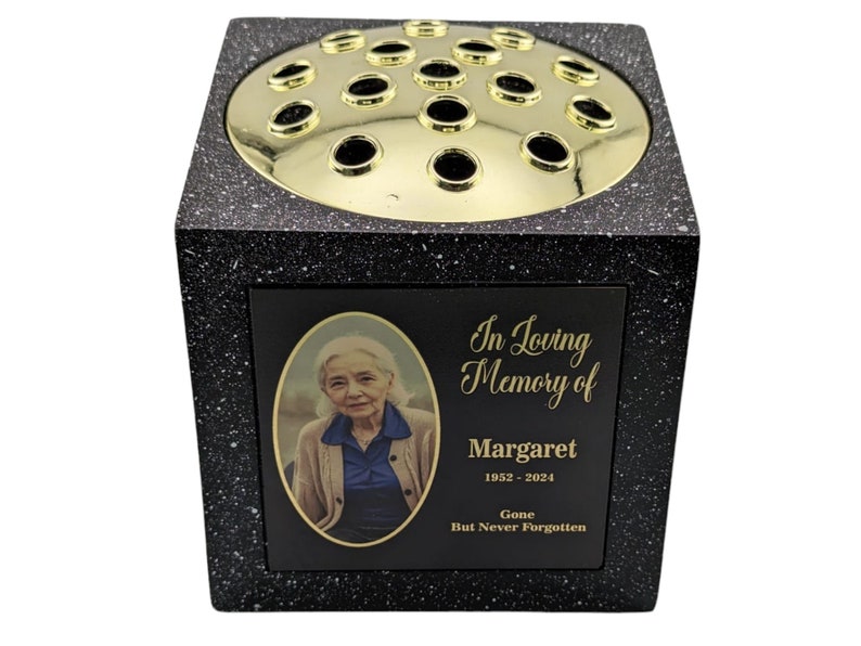 May include: A black and speckled memorial vase with a gold-colored floral grid. The vase features a photo of a woman and the inscription "In Loving Memory of Margaret 1952-2024 Gone But Never Forgotten."