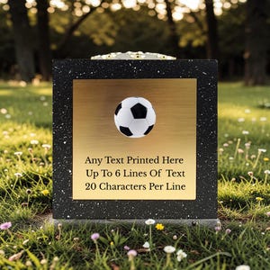 Op de afbeelding: Een zwart en goud voetbal-thema gedenkplaat. De plaat heeft een zwart gespikkelde rand en een gouden midden met een voetbal. Tekst: "Any Text Printed Here Up To 6 Lines Of Text 20 Characters Per Line."
