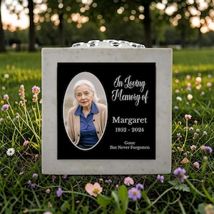 May include: A memorial stone with a black plaque featuring a photo of a woman in an oval frame. The text reads "In Loving Memory of Margaret 1952-2024 Gone But Never Forgotten." The stone is light grey and sits on grass.