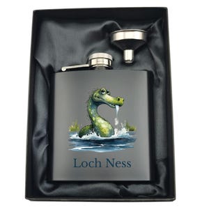May include: A stainless steel flask with a black background and a cartoon illustration of the Loch Ness Monster. The flask has a silver funnel and the text "Loch Ness" printed on the front.