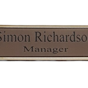 May include: A gold-framed name tag with a brown background. The name tag reads "Simon Richardson, Manager."