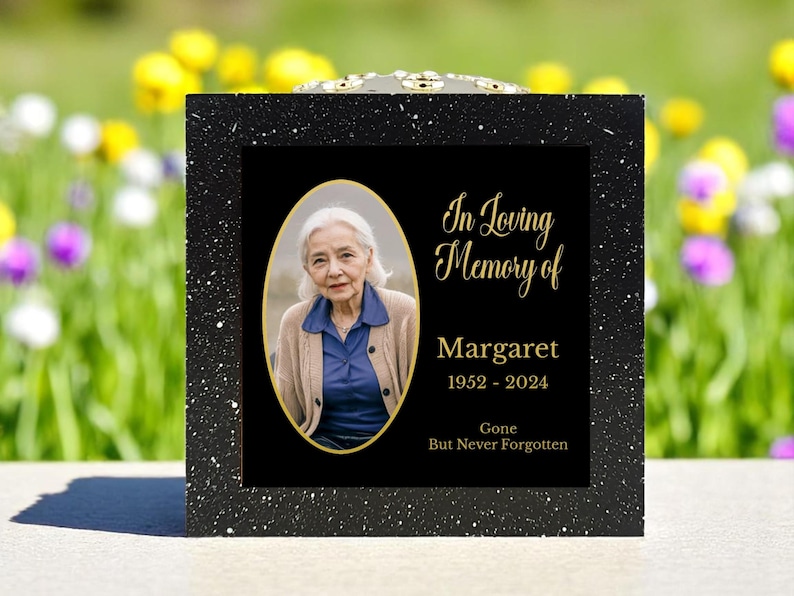 May include: A black memorial box with a speckled finish. The front features a photo of a woman in an oval frame, with the text "In Loving Memory of Margaret 1952-2024 Gone But Never Forgotten" in gold lettering. A remembrance item.