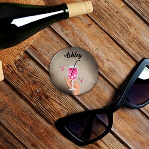 Puede incluir: Un posavasos redondo con un vaso de cóctel rosa y el nombre Ashley impreso en él. El posavasos está sobre una superficie de madera con una botella de vino y gafas de sol.