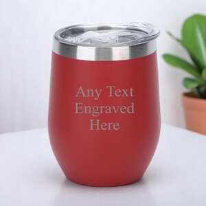 May include: A red insulated tumbler with a silver rim and a clear lid. The tumbler has the text "Any Text Engraved Here" in a light grey colour. A small potted plant is in the background.