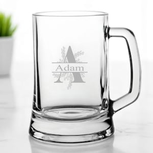 Könnte beinhalten: Klarer Bierkrug mit Henkel, mit dem Namen "Adam" in dekorativer Schrift mit floralen Akzenten graviert. Der Krug ist leer und steht auf einer weißen Oberfläche. Eine kleine Topfpflanze befindet sich im Hintergrund.