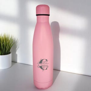 以下が含まれることがあります： 黒色の蓋と花柄のデザインが施されたピンク色のステンレス製水筒。側面には「C」の文字と「Claire」という名前が刻まれています。