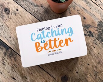 May include: A white rectangular bait tin with the text "Fishing is Fun, Catching is Better" in blue and orange. The tin also has three small fish illustrations and the text "John's Bait Tin". A small potted succulent sits in the background.