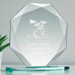 May include: A clear glass golf-themed award with an octagonal shape. The award features an engraved golf ball, flag, and clubs design. The text "Any Text Engraved Here" is also visible. A small potted plant is in the background.