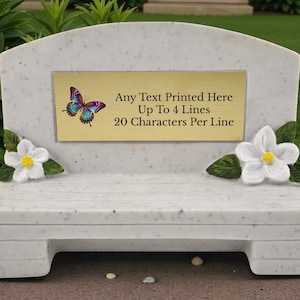 Op de afbeelding: Een witte marmeren bank met een gebogen rugleuning en een rechthoekige plaat. De plaat toont een vlinder en de tekst "Any Text Printed Here Up To 4 Lines 20 Characters Per Line". Witte bloemen en groene bladeren sieren de zijkanten.
